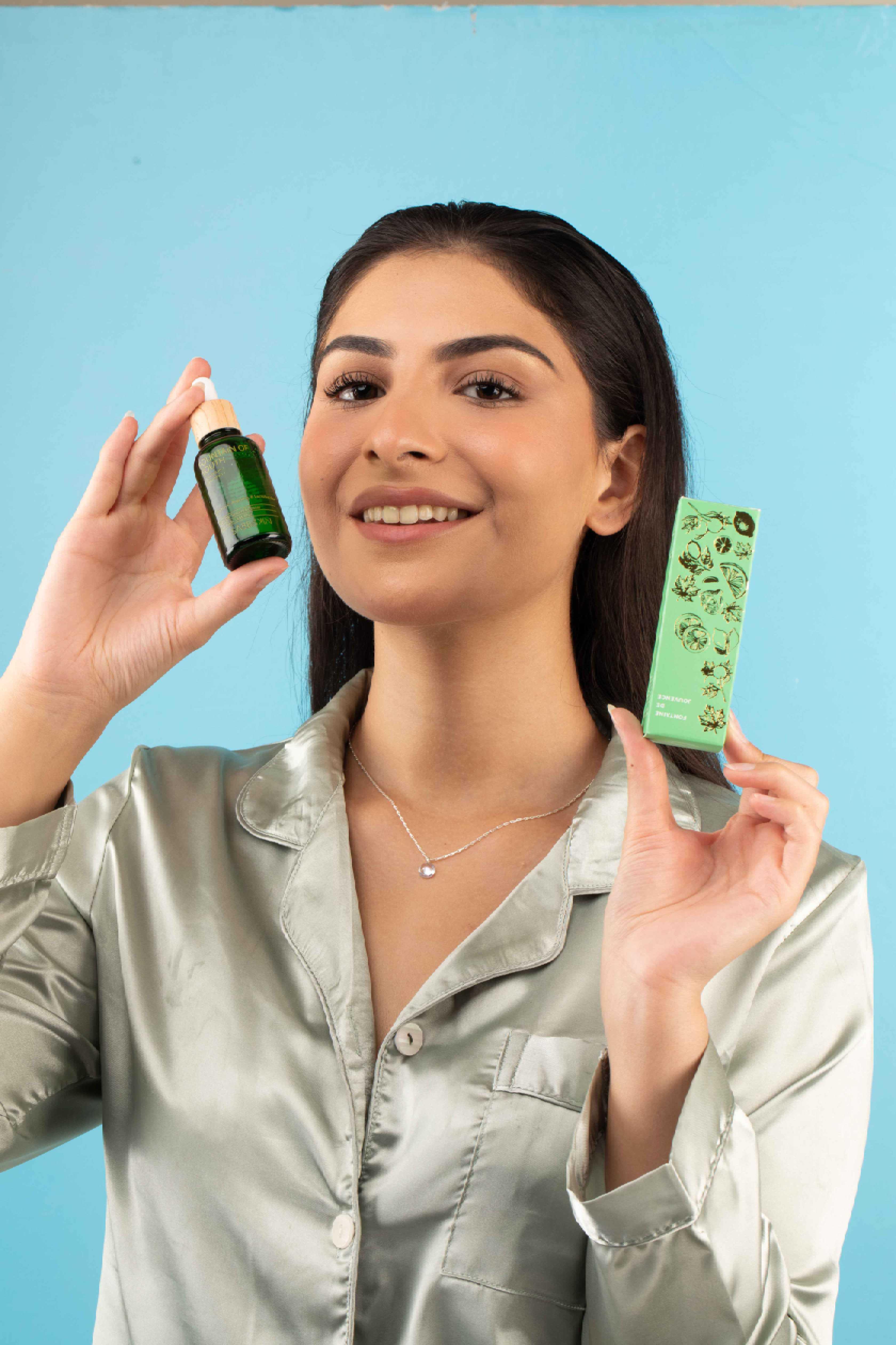 Smiling woman in silk shirt holding green serum bottle and matching skincare box against blue background